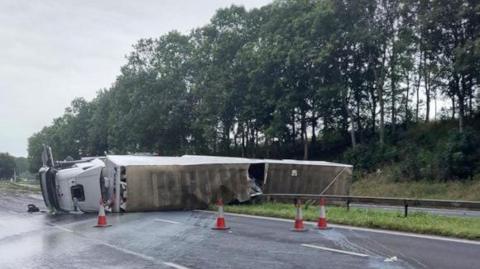 The lorry, which has a white cab and trailer, turned on its side across the central reservation of the M6. A number of traffic cones are placed around the damaged vehicle.