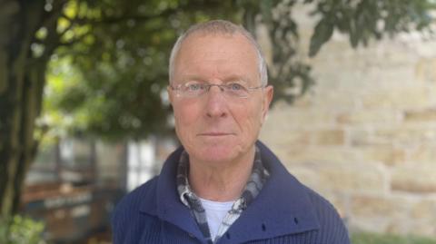 Chris Weatherley stands outside, with hedges and a stone building in the background. He is wearing glasses and a blue jumper. 