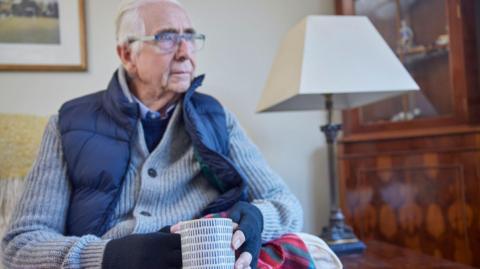 An elderly man with grey hair and glasses in his home.  He is wearing four layers of clothing and fingerless gloves.  He is holding a mug and using a blanket over his knees. 
