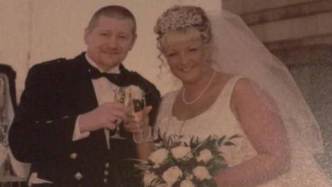 A man in traditional Scottish dress including a black bow-tie, waistcoat and kilt, stands next to a woman in a white wedding dress and veil. Both are smiling and are clinking glasses of champagne.