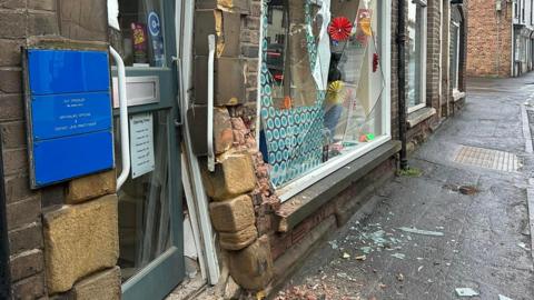 A shop's front window has been completely smashed and the brickwork surrounding it has also been damaged. There are shards of glass and debris on the footpath in front of it. 