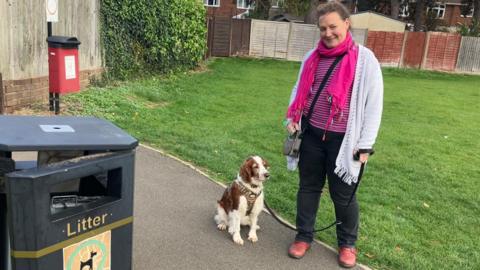 Paula is seen wearing a pink scarf and light cardigan stands on a park path holding her dog’s lead beside a litter bin. The brown-and-white spaniel sits at her feet.