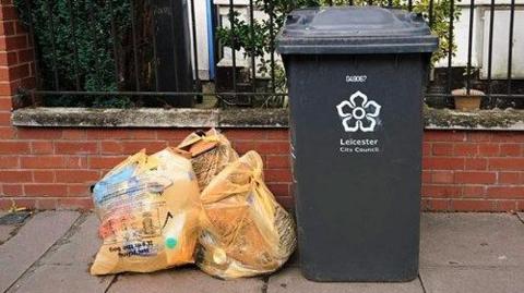 Three orange recycling bags on a pavement next to a black bin with a Leicester council crest