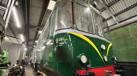 The front of the railbus is dark shiny green with yellow stripes pointing downwards in the middle. A bright red buffer and several lights. It is inside a warehouse surrounded by equipment.