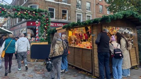 A busy outdoor Christmas market with decorated wooden stalls, shoppers and a brick building with a Pret A Manger in the background.