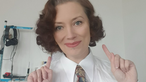 A woman with curly bobbed hair points one finger on each hand towards herself. She is wearing a shirt and tie and is smiling at the camera

