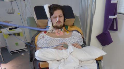 Nick Atherton sitting in a chair on a hospital ward. He has a tube coming out of his nose, one attached to his hand and his head and neck are supported by a variety of equipment. He is wearing a pale hospital gown and has a dark beard and short dark hair. His arms are resting on white pillows on his lap
