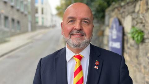 A bald man with a grey beard and moustache. He's wearing a blue suit and a white shirt with a bright red and yellow striped tie. 