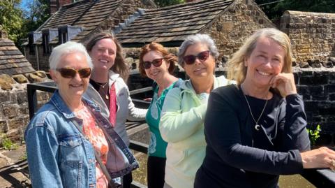 Five women stand along a barrier with a canal behind them. They are all dressed casually in jumpers and smile at the camera.