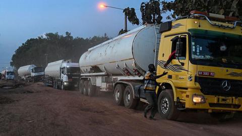 A line of large Malian fuel tankers stretches along a dusty road at dawn or dusk, their headlights and a streetlamp glowing through a hazy sky. A man walks beside the first yellow truck, holding on to its door as the convoy waits.