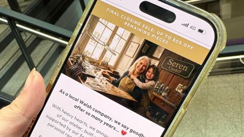 A mobile phone being held in someone's hand. On the phone is an image of an older woman with an apron sitting at a workshop desk, with a young girl hugging her. A sign saying 'Seren Cardiff' can be seen in the background. 
