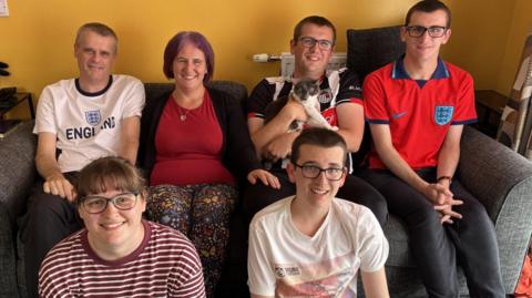 A family of six are sitting in a living room with yellow walls. There are three men and one woman on a grey sofa and one of the men is cuddling a grey and white cat. Sitting on a grey carpet in front of the sofa is another woman and a teenage boy of 16. Everyone is smiling