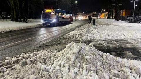 SNow piled up by the side of a cleared road as a bus passes