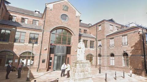 Exterior of Carlisle Crown Court. It is a three-storey red bricked building with a statue outside the front entrance. There are anonymous people standing outside (stock image).