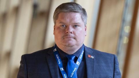 Chris McEleny, a fair haired man wearing a blue suit shirt and tie, photographed in the Scottish Parliament. He is visible from the chest up. 