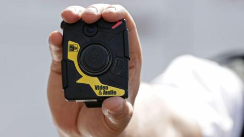 The outstretched arm of a person holding a black and yellow body-worn camera.