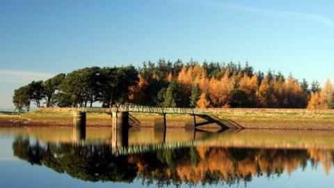 The dam at Harlaw reservoir. There is a jetty jutting out into the water and trees in the background. There is a reflection of the trees on the water.