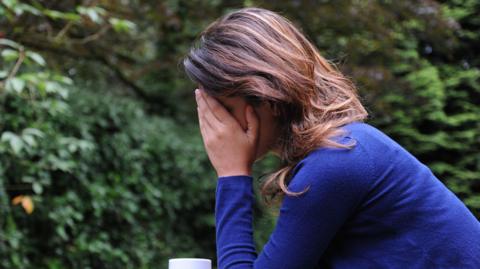 A woman with long brown hair sits in a garden with a mug in one hand while her other hand is raised to her head and covers her face. She is wearing a blue cardigan with blue jeans.