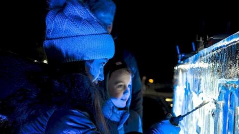 A girl in a black hooded coat and woolly hat marking something into an ice sculpture in front of her with another child stood next to her smiling