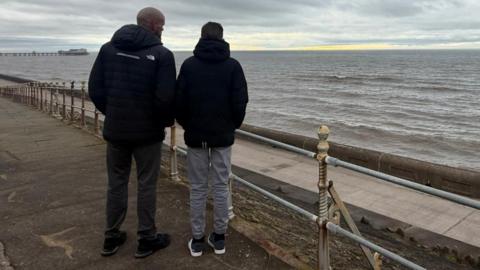A man wearing a black North face jacket stands next to a teenager with grey trousers looking out to sea on a grey day. 