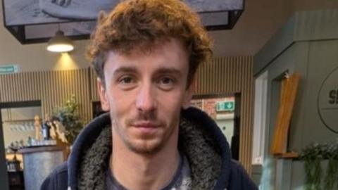The image shows a man with light brown untidy hair and facial stubble on a thin white face. He is seated at a table in a café. He is wearing a dark zip‑up hoodie over a patterned T‑shirt.