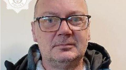 A mugshot of a bald man with rectangular glasses. He is staring into the camera and has stubble around his chin and upper lip. The hood of a dark grey jumper can be seen at the bottom of the image.