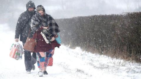A cold-looking man and woman walk along a snow-covered path beside a hedge in a blizzard. They are wearing winter coats and carrying groceries including toilet roll.