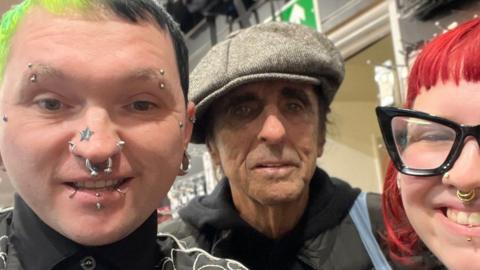 Adhum Price, a man with bright green and black hair and numerous facial piercings, is smiling while posing for a selfie alongside Alice Cooper who is wearing a tweed flat cap and stood in between Mr Price and a store colleague who has bright red hair and is wearing black, thick-rimmed glasses.