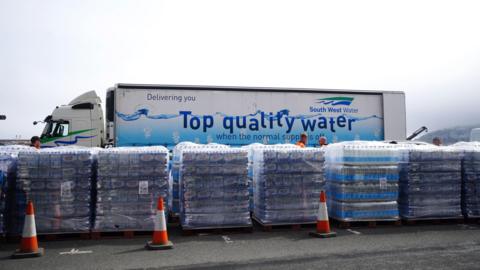 Six big packages of bottled water in a car park. Behind them is a lorry with South West Water branding and the line: Delivering you top quality water.