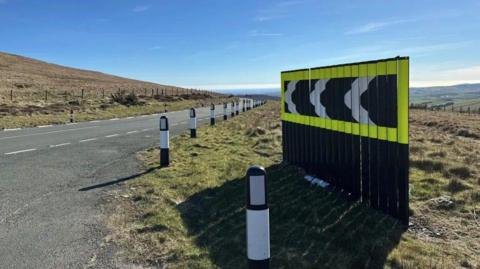 A board of black and white chevrons bordered in bright yellow at the side of the Mountain Road on sunny day with blue sky above.