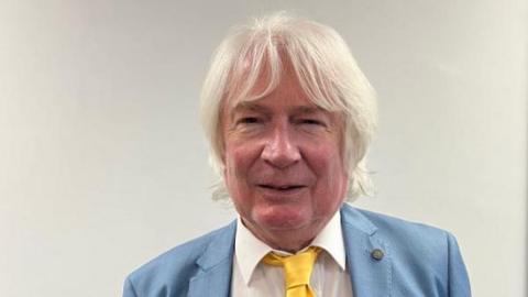 A man with collar length grey hair is looking at the camera. He is wearing a pale blue suit, white shirt and yellow tie