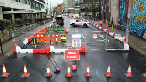 A road that has been sealed off with fencing and traffic cones, as well as a Road Closed sign. Workers in orange high vis jackets can be seen in the background.