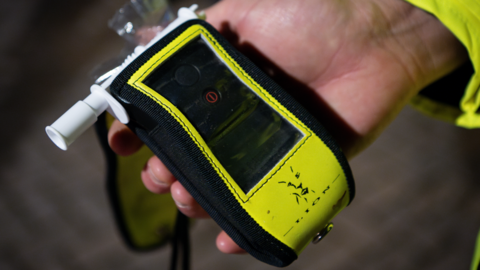 A close up of a breathalyser being held in a police officer's hand. It is a slightly bigger than a mobile phone and has a white tube for drivers to blow into. It is in a yellow and black protective case.