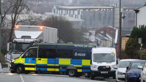 A blue and yellow police van is parked across the road , which is lined with vehicles. Bolton's skyline is in the background.