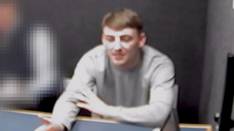 Kane Farragher is in a grey police interview room and is speaking with someone off-camera with one arm outstretched and the other crossed over his body. He has short brown hair and is wearing a grey jumper. There is someone in blue sitting beside him, but they are blurred out.