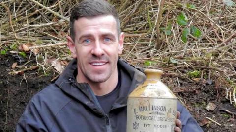 A person sitting in a muddy digging site, holding a ceramic bottle they had just unearthed. The bottle shows printed text from a botanical brewery.