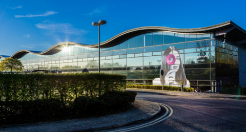 A large, glass building with a wavy roof. There is a graphic of a woman peering through one of Dyson's ring hairdryers stuck on to the right-hand side. A road with a hedge leads up to the building. 