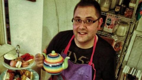 An old photograph of Kevin Sargeant who has dark hair and glasses and is wearing a black t-shirt and a purple apron. He is holding up a stripey teapot next to an afternoon tea stand which has a range of cakes on it