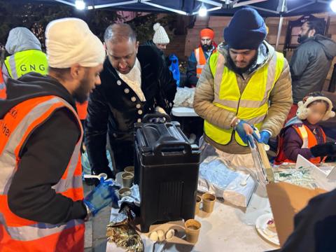 A team of Sikhs volunteer at the outdoor soup kitchen, wearing high vis vests and blue plastic hygiene gloves. Two of the men wear a turban, one white and one blue. There is a table in front of them with cups of coffee and a large black box, presumably a coffee machine. They stand under a gazebo with lights hanging from it. It is night time 