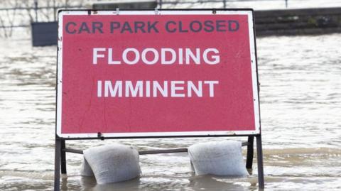 A sign with a red background with Car park closed writted in black and flooding imminent written in whit. There are two sandbags weighting it down and water surrounding it.