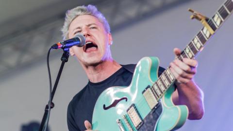 Callum Beattie has styled blonde hair and wearing a black T-shirt. He is standing on a stage and is singing into a microphone and holding an aquamarine coloured electric guitar.