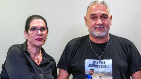 Talik, who has black hair in a ponytail and wears black-rimmed glasses, poses for a photo with her husband Itzik, who has grey short hair and a beard and wears a silver chain. They both have neutral expressions. 
