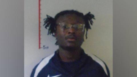 A mugshot depicting man with dark hair and large glasses, he is wearing a navy tracksuit top.