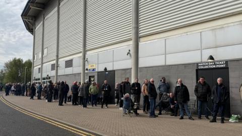 A long queue of people outside a sports stadium. They are waiting near a sign which reads “Match Day Ticket Collection Point". They are standing on a paved walkway next to a tarmac road under an overcast sky.