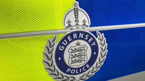 The side of a yellow and blue police vehicle. At the centre is a Guernsey Police badge which is silver and dark blue.