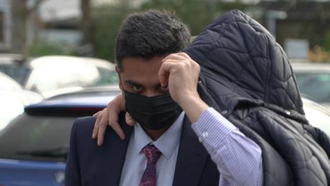 Salil Korambayil wearing a black face mask and being partly shielded by a man with a hood pulled up