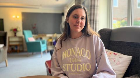 Jess is wearing a pink jumper. She has long brown hair and is looking into the camera.