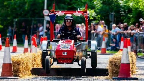 A man in a helmet and white sunglasses is in a homemade soapbox kart which has smaller black wheels at the front and bigger ones at the back. It has a red front frame and red frame above his head. It is surrounded by cones and hay bales with spectators behind a railing in the distance.