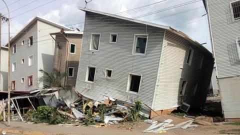 A damaged multi-storey residential building leaning to one side, with broken windows, loose cladding and debris scattered around its base.