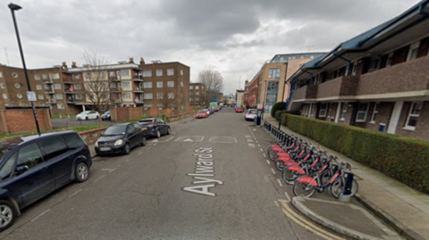 A street view of Aylward Street in Stepney Green, east London, showing parked cars on both sides of a residential road under a cloudy sky. A row of docked rental bicycles lines the pavement on the right, with low-rise brick apartment buildings on either side of the street.